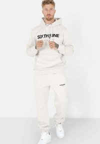 Lys beige hettegenser med svart "SIXTH JUNE" logo, parret med matchende joggebukser. Hettegenseren har kengurulomme; joggebuksene har sidelommer.