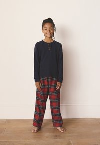 Navy long-sleeve shirt with three brown buttons, paired with red plaid pants featuring blue and green accents. Model stands barefoot on wood floor.