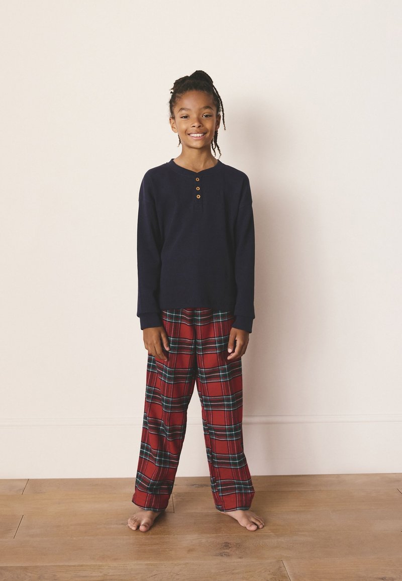 Soe ja pruunide nuppudega tumesinine pikkade varrukatega särk, millele on kombineeritud punased ruudulised püksid siniste ja roheliste lisanditega. Modell seisab paljajalu puidust põrandal.