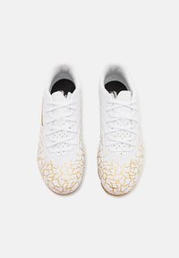Des chaussures de sport blanches présentent une tige texturée avec un motif doré craqué distinctif, des lacets plats et un design à profil bas avec des perforations.