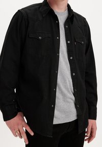 Musta denim-paita, jossa on nepparit, kaksi etutaskua ja klassinen kaulus. Paita on pukeutuneena vaaleanharmaan T-paidan päälle, jossa on röpelöinen pinta.