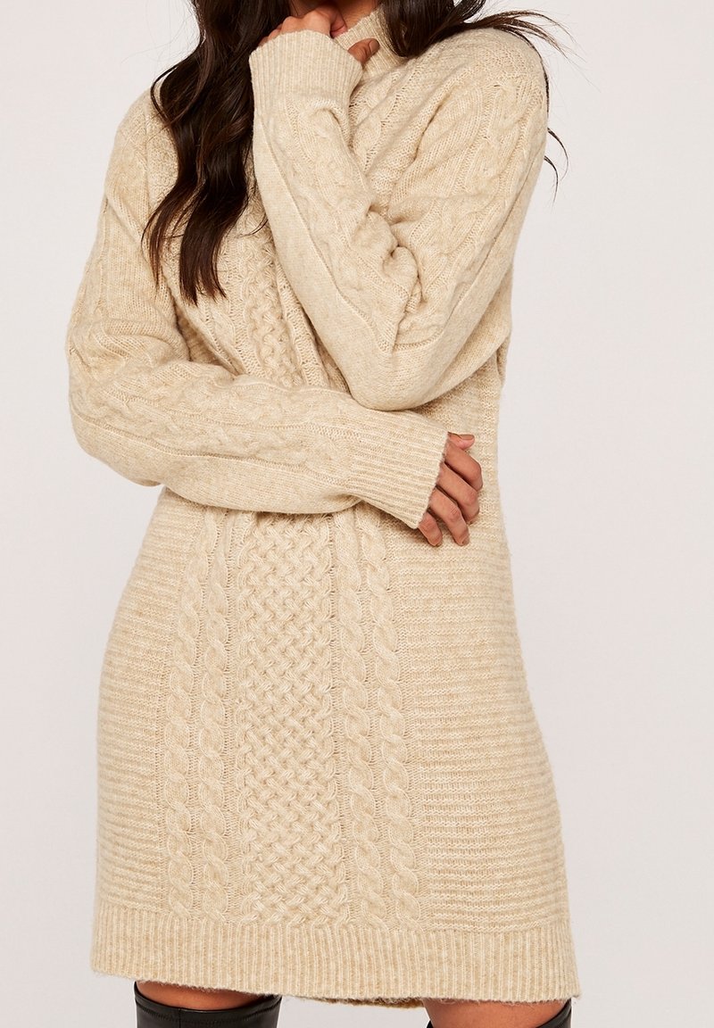 Femme portant une robe-pull beige en maille torsadée à manches longues, bras croisés, et des bottes noires montantes, photographiée sur un fond uni.