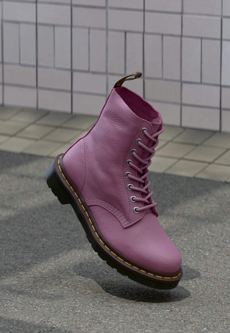 Une botte en cuir rose à lacets avec une semelle noire et des coutures jaunes sur un trottoir gris près d’un mur carrelé.
