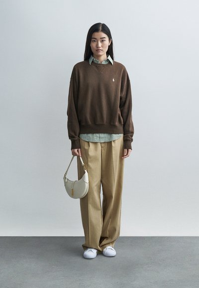 Rjav pulover z logotipom, nošen čez zeleno-belo črtasto srajco. Bež hlače širokih nog, bele čevlje in majhna kremna torbica.