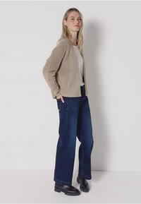 Beige, texturerad kofta över en vit topp, tillsammans med mörkblå vida jeans och svarta ankelboots. Enkelt, avslappnat passformdesign.