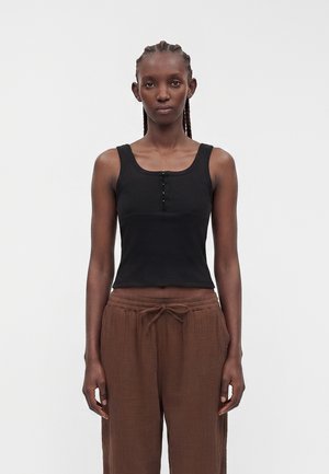 Jeune personne aux cheveux tressés portant un haut noir sans manches avec des boutons et un pantalon marron ample, debout devant un fond clair uni.