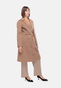 Cappotto di lana color beige con design avvolgente, maniche lunghe e cintura. Presenta tasche laterali e una texture liscia, abbinato a pantaloni beige e tacchi neri.