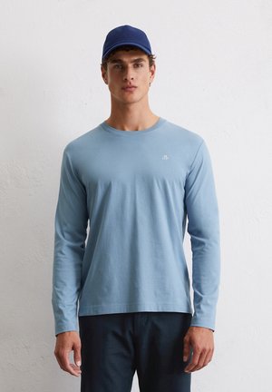 Lichtblauw shirt met lange mouwen van zachte stof en een ronde hals, gecombineerd met een blauwe pet. Minimalistisch logo op de borst. Donkere broek.