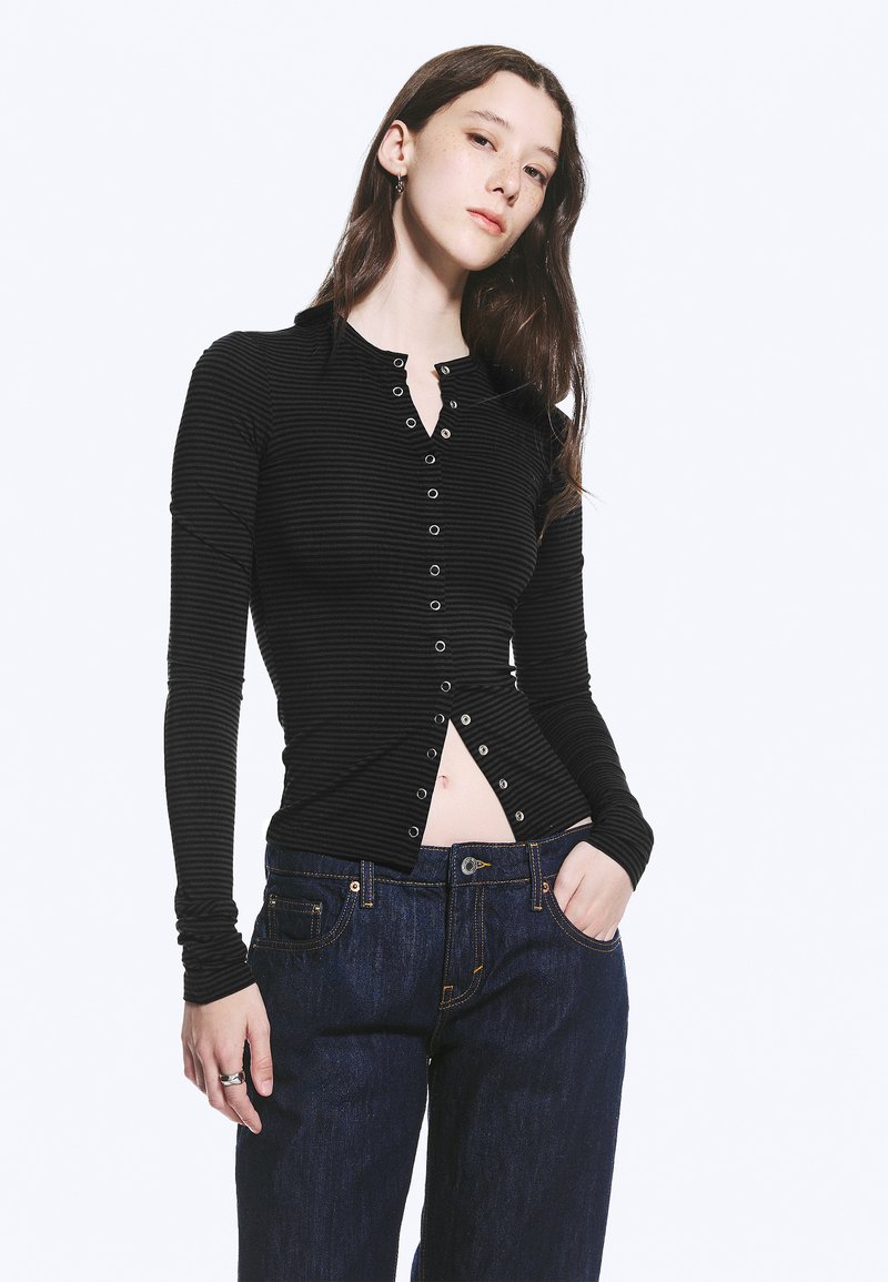 Jeune femme aux longs cheveux bruns portant un haut noir ajusté à boutons et un jean bleu foncé, debout avec une main dans la poche.