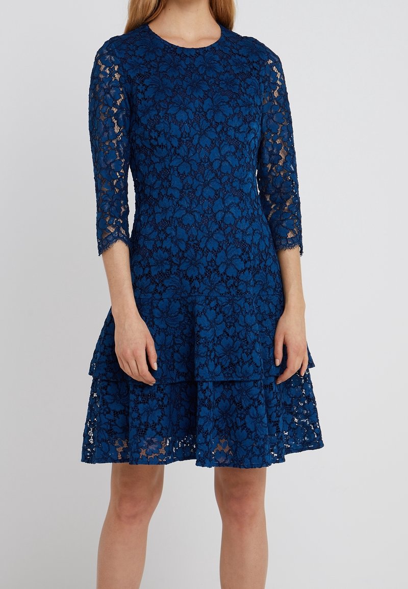 Femme portant une robe en dentelle bleue avec des manches trois-quarts et une jupe à étages, debout devant un fond uni gris clair.