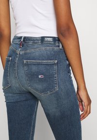 Tmavě modré džíny vyrobené z denimu s mírně vybledlým povrchem, s etiketou Tommy Jeans a červenobílomodrým logem na zadní kapse.