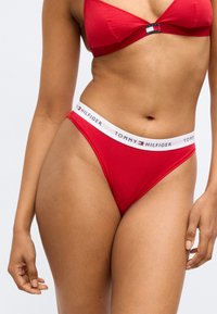 Rødt bikinitøj af blødt stof, bestående af en bralette med trekantede cups og trussebund i g-streng-stil med en hvid, mærket talje.