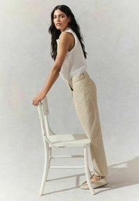 Haut sans manches blanc avec des bords festonnés, pantalon taille haute beige clair, et chaussures tressées beige. S'appuyant sur une chaise en métal blanc.