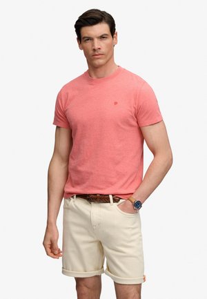 Man draagt een koraalkleurig t-shirt met ronde hals, beige opgerolde korte broek, bruine gevlochten riem en polshorloge, staat met één hand in de zak.