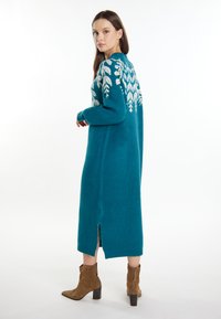 Cardigan lung teal, tricotat, cu motive albe în formă de frunză pe spate, având o deschidere laterală și mănici cu bordură. Modelul poartă botine negre.
