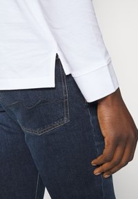 Weißes Langarmhemd mit seitlichen Schlitzen, kombiniert mit dunkel gewaschenen Jeans, die ein dezentes Taschen-Design und Nähdetails aufweisen.