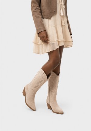 Kvinde iført beige ruskind cowboystøvler med detaljeret syning, kombineret med et lysebeige lagdelt skørt og en brun jakke.
