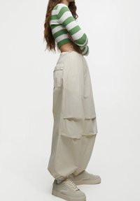 Beige Cargo-Hosen mit mehreren Taschen, lockerer Passform und gerafften Knöchelbündchen, kombiniert mit hellgrauen Sneakers und einem grün-weiß gestreiften Oberteil.
