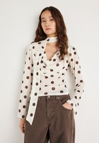 Kremowa bluzka z długim rękawem w brązowe groszki z dekoltem typu komin. Zestaw uzupełnia dopasowany szalik i miękka, teksturowana tkanina.