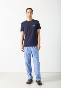 Vyras stovi atsipalaidavęs rankas, dėvėdamas tamsiai mėlyną marškinėlį, mėlynas ir baltas dryžuotas kelnes bei juodus klumpes ant vienspalvio balto fono.