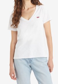 White cotton t-shirt with a V-neck and short sleeves. Features a small red logo on the chest. Paired with light blue denim jeans.