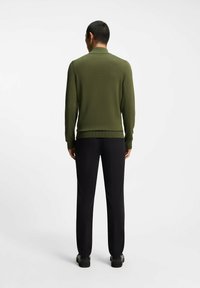 Maglione verde oliva con collo e polsini a coste, abbinato a pantaloni neri. Il tessuto appare liscio e aderente su tutta la superficie.