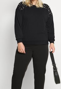 Svart sweatshirt med utsmyckad axeldetalj, rund halsringning och ribbade ärmslut, tillsammans med svarta byxor och en svart handväska.