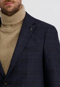 Mann trägt einen beigen Rollkragenpullover und ein dunkelblaues Karoblazer mit einem auf der linken Seite angebrachten Anstecker in Form des Eiffelturms.