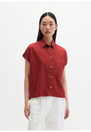 Femme aux cheveux noirs et raides portant une chemise rouge à manches courtes boutonnée et un pantalon blanc taille haute, debout devant un fond blanc uni.