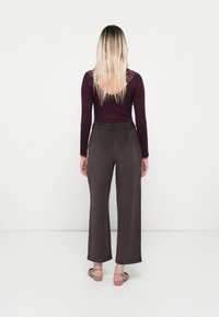 Femme aux longs cheveux blonds portant un haut à manches longues violet foncé avec de la dentelle, un pantalon large marron foncé et des sandales beiges, debout face au mur.