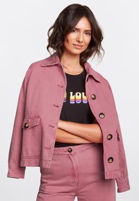 Rosa denimjacka med knappstängning, bröstficka och krage. Bärs över en svart t-shirt med färgglad text.