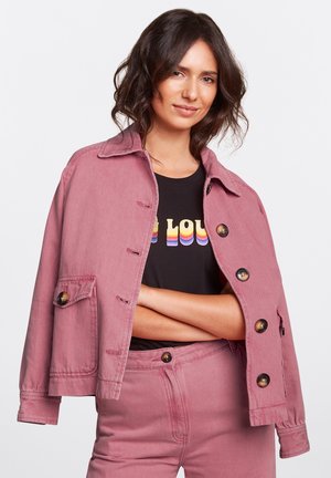 Veste en denim rose avec fermeture à boutons, poche poitrine et col. Portée par-dessus un t-shirt noir avec des lettres colorées.
