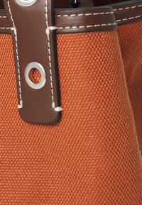 Sac en tissu orange tissé avec des accents en cuir marron, doté de quincaillerie en argent. Gros plan montrant la texture et les détails de couture de la sangle.