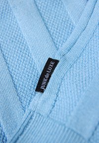 Hellblaues Strickkleidungsstück mit einem kleinen schwarzen Etikett mit der Aufschrift "JUNK de LUXE", das an einer Naht in der Nähe des V-förmigen Halsausschnitts eingenäht ist.