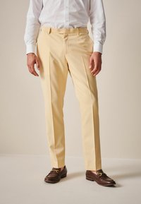 Beige maßgeschneiderte Hose aus glattem Stoff mit vorderen Falten und Knopfverschluss, kombiniert mit dunklen Slippern mit goldenen Akzenten.