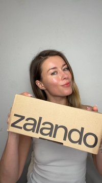 Kvinne med langt hår holder en brun pappeske med "zalando" trykt i svart, smilende svakt mot en enkel lys grå bakgrunn.