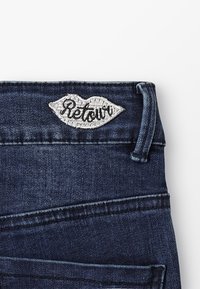 Vaqueros de mezclilla oscura con un logo bordado en forma de labios que dice "Retouri". Detalles de costura distintivos y pretina funcional.