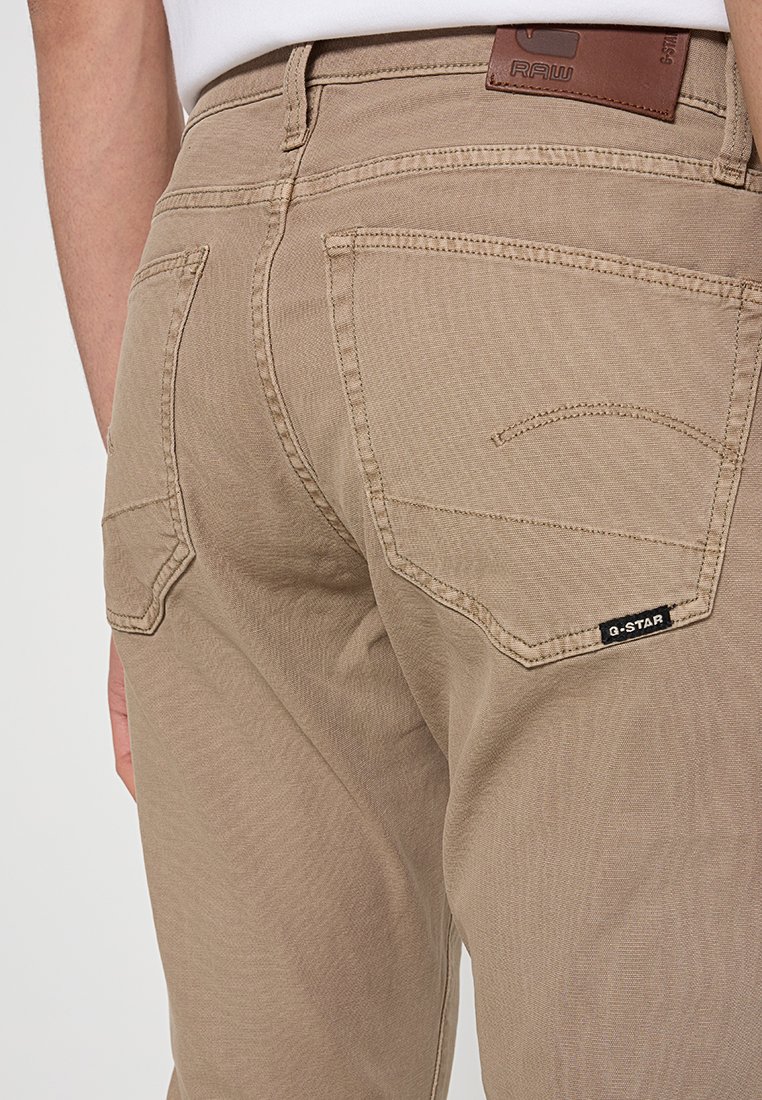 Beżowe bawełniane spodnie z skórzanym naszywką z napisem 'RAW' oraz metką logo. Tylnie kieszenie mają subtelne wzory przeszyć oraz zaokrąglone brzegi.