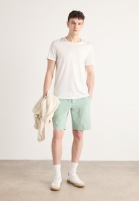 Hvid t-shirt med korte ærmer, mintgrønne shorts, hvide sneakers med lysebrune skoletsåler, holder en beige jakke. En simpel, afslappet påklædning.