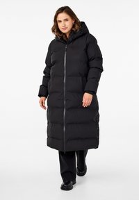 Manteau en duvet noir avec capuche, présentant une fermeture éclair à l'avant, une fermeture à bouton sur le côté et une texture matelassée. Porté avec un pantalon noir et des bottes.