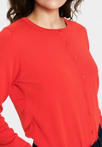 Červený top s dlouhým rukávem a kulatým výstřihem, s knoflíkovou légou a vroubkovanou strukturou kolem límečku a manžet.