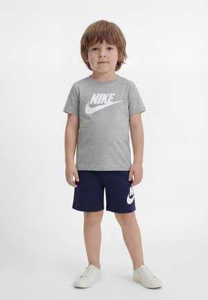 Młody chłopiec stoi przodem, ubrany w szarą koszulkę Nike, granatowe spodenki Nike i białe trampki na białym tle.