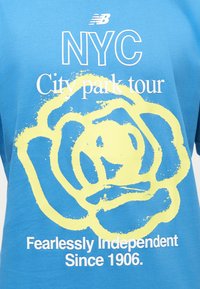 Μπλε μπλούζα New Balance με το κείμενο «NYC City park tour», μια κίτρινη αφηρημένη ανθοδέσμη και το κείμενο «Fearlessly Independent Since 1906» μπροστά.