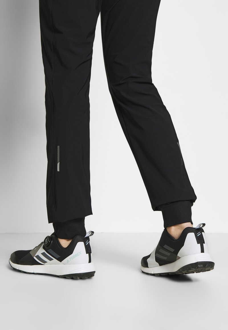 Person, der schwarze Sporthosen und schwarze und weiße Adidas Terrex Sneakers trägt, steht auf einem weißen Boden vor einem schlichten Hintergrund.
