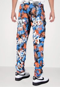 Abstrakte Cargo-Hosen in einem lebhaften Blau-, Orange- und Schwarzspritz-Muster, mit Taschen und weißen grafischen Textelementen.