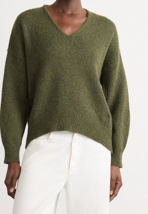 Osoba v olivově zeleném svetru s výstřihem do V, s žebrovanými manžetami a lemem, v kombinaci s bílé kalhoty s vysokým pasem na jednobarevném pozadí.