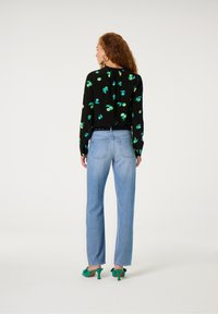 Schwarze Bluse mit grünem Blattmuster, blaue gerade Jeans und grüne High-Heels mit Mule-Design. Lange Ärmel und lockere Passform, glatte Textur.