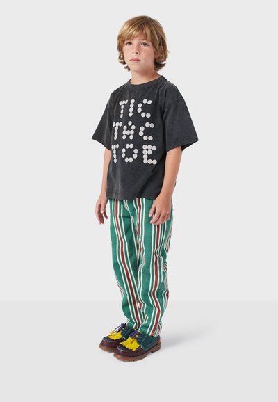 Zwarte oversized t-shirt met witte "TIC TAC TOE" graphic, groene en rode gestreepte broek, multicolored schoenen met franjes en hardware accenten.