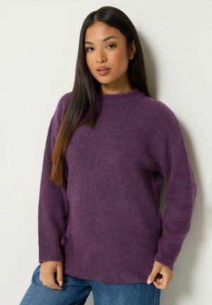 Kvinde med langt mørkt hår iført en lilla strikket sweater og blå jeans, der står mod en hvid væg.