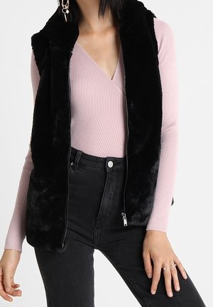 Vrouw draagt een zwarte mouwloze faux fur bodywarmer over een roze geribd wikkel-topje en zwarte high-waisted jeans.